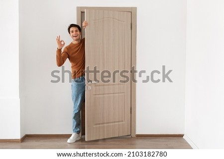 Good Offer. Cheerful young guy standing in doorway of his apartment, smiling millennial male homeowner holding ajar door looking peeping out greeting visitor, showing ok sign gesture, full body length