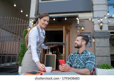 Good service. Smiling waitress bringing coffee to bearded male with smartphone - Powered by Shutterstock