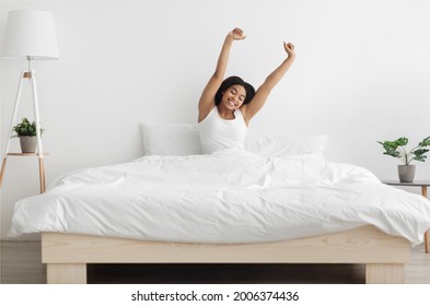 Good start of new day. Well-slept african american lady stretching arms after waking up, feeling happy and full of vitality. Young woman sitting in comfortable bed in coy bedroom interior - Shutterstock ID 2006374436