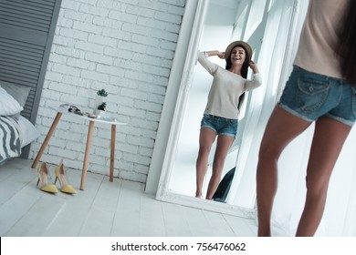 
So good to stay at home. Rear view of beautiful young woman fitting hat and looking at her reflection in the mirror with smile while standing indoors.
