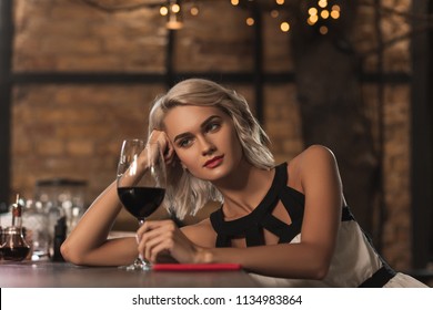 Good wine. Beautiful young blonde woman sitting at the bar counter and looking at her glass of wine while thinking about something