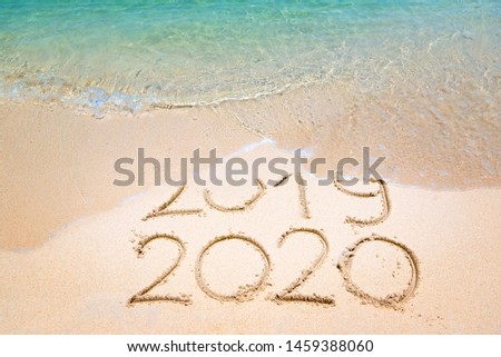 Goodbye 2019 Happy New Year 2020 lettering on beach with wave and clear blue sea on sunny day. Handwritten inscription 2019 and 2020 on beautiful golden sand beach. New Years 2020 replace 2019 concept