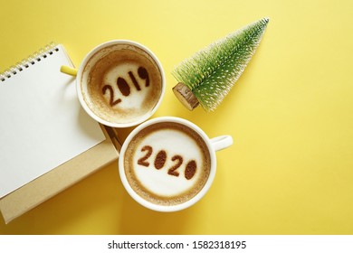 Goodbye 2019 Hello 2020 Holidays Food Art Theme Coffee Cup With Number 2020 On Frothy Surface, Another One With 2019 At The Bottom Of Cup Over Yellow Background With Christmas Tree And Blank Calendar.