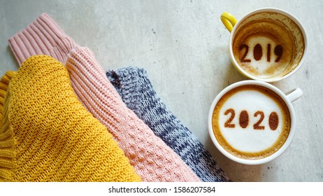 Goodbye 2019 Hello 2020 Holidays Food Art Theme Coffee Cups With Number 2020 And 2019 Over Frothy Surface On Grey Cement Background With Knitted Long Sleeves Sweaters. (top View, Space For Text)