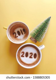 Goodbye 2019 Hello 2020 Holidays Food Art Theme Coffee Cup With Number 2020 On Frothy Surface, Another One With 2019 At The Bottom Of Cup Over Yellow Background With Christmas Tree. (space For Text)