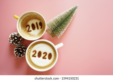 Goodbye 2019 Hello 2020 Holidays Food Art Theme Coffee Cup With Number 2020 On Frothy Surface, Another One With 2019 At The Bottom Of Cup Over Pink Background With Christmas Tree And Pinecones.