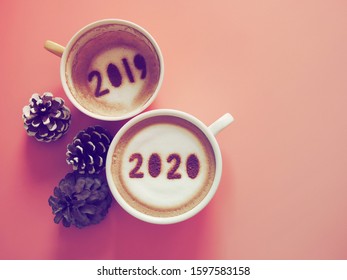 Goodbye 2019 Hello 2020 Holidays Food Art Theme Coffee Cup With Number 2020 On Frothy Surface, Another One With 2019 At The Bottom Of Cup Over Pink Background With Pinecones. (space For Text)