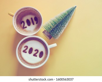 Goodbye 2019 Hello 2020 Holidays Food Art Theme Coffee Cup With Number 2020 On Frothy Surface, Another One With 2019 At The Bottom Of Cup Over Yellow Background With Christmas Tree. (space For Text)