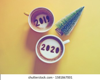 Goodbye 2019, Hello 2020 Theme Coffee Cup With Number 2020 On Frothy Surface And Another One With 2019 At The Bottom Of Cup Over Yellow Background With Christmas Tree. Holidays Food Art Concept.