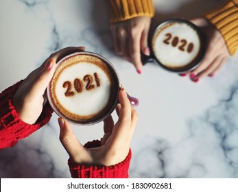 Goodbye 2020, Hello 2021 Theme Coffee Cup With Number 2021 On Frothy Surface In Female Hands Holding And Another One With Number 2020 On Frothy Surface Over Marble Table Background. Holidays Food Art.