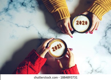 Goodbye 2020, Hello 2021 Theme Coffee Cup With Number 2021 On Frothy Surface In Female Hands Holding And Another One With Number 2020 On Frothy Surface Over Marble Table Background. Holidays Food Art.