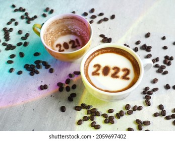 Goodbye 2021, Hello 2022 Theme Coffee Cups With Number 2022 And 2021 Over Frothy Surface On White Cement Background With Coffee Beans And Rainbow Lights. Holidays Food Art For Happy New Year.