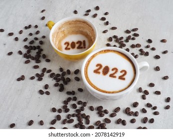 Goodbye 2021, Hello 2022 Theme Coffee Cup With Number 2022 On Frothy Surface And 2021 At Bottom Of Another One On Cement Background With Coffee Beans. Holidays Food Art Concept For Happy New Year.