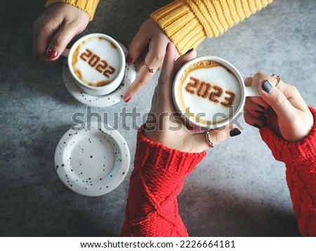 Goodbye 2022, Hello 2023 coffee cups with number 2023 and 2022 over frothy surface in female hands holding on dark table background. Happy New Year holidays food art theme. (selective focus on 2023)