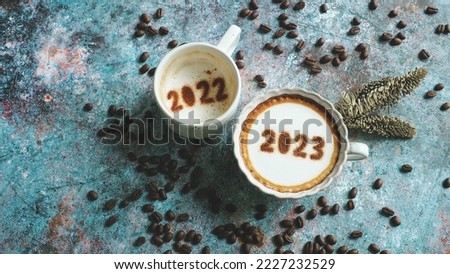 Goodbye 2022, Hello 2023 theme coffee cups with number 2023 and 2022 over frothy surface flat lay on rustic blue background with coffee beans and dried pine branches. Holidays food art Happy New Year.