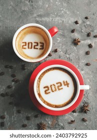 Goodbye 2023 Hello 2024 holidays food art theme coffee cup with number 2024 on frothy surface, another one with 2023 at the bottom over dark rustic grey background with coffee beans and star anise.