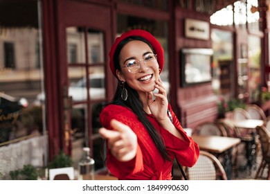 Good-humored Asian tanned woman in red beret, stylish dress and eyeglasses smiles and pulls hand to camera. Pretty brunette girl poses in street cafe.