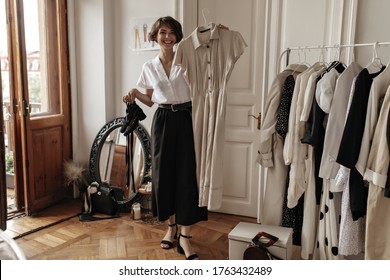 Good-humored stylish young woman in midi skirt, white blouse smiles sincerely, holds beige linen dress and black shoes, poses in dressing room.