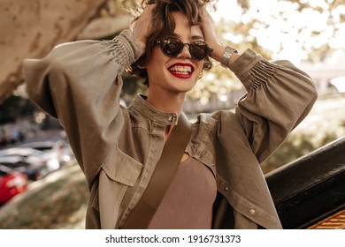 Good-humored Woman With Brunette Hair And Red Lips Laughing Outside. Modern Girl In Denim Jacket And Glasses Smiling Outdoors..