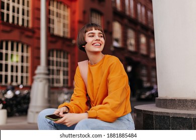 Good-humored woman sits outside. Short-haired girl in orange sweater holds smartphone and smiles outdoors. - Powered by Shutterstock