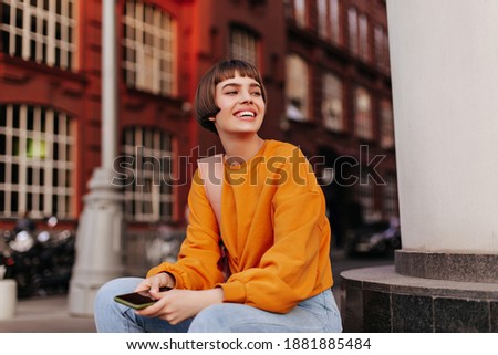 Good-humored woman sits outside. Short-haired girl in orange sweater holds smartphone and smiles outdoors.