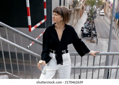 Good-humored Woman Standing Outside. Short-haired Girl In Black Shirt And White Pants In Wireless Headphones Holding A Bottle Of Water And Smiles Outdoors.