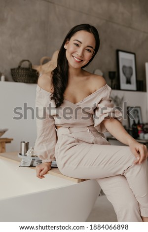 Good-humored woman in stylish beige pants and linen blouse smiles widely, looks into camera and sits on bath in cozy bathroom.