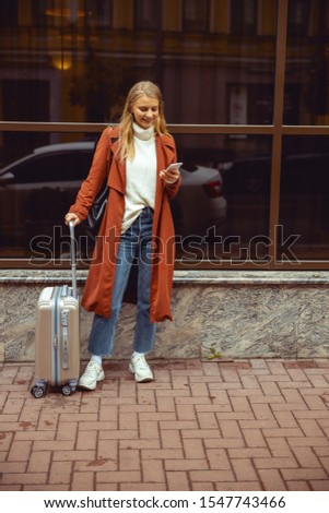 Good-humored young female tourist leaning on her trolley suitcase outdoors