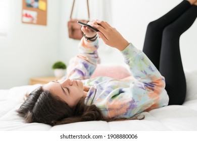 Good-looking Adolescent Girl Lying In Bed With Her Feet Up In The Wall. Caucasian Teen Girl Scrolling Through Social Media And Texting On Her Smart Phone