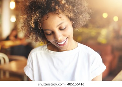 Good-looking African Woman With Afro Haircut Dressed In White Top, Enjoying Day-off At Cafeteria, Looking Down With Charming Shy Smile. Black Girl Sitting At Table Against Blurred Interior Background