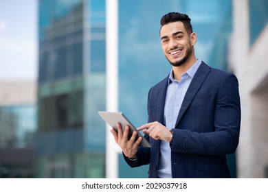 Good-looking arab guy in formal wear standing by office building, holding digital tablet and smiling, looking for new business opportunities online, using business application, copy space, panorama - Shutterstock ID 2029037918
