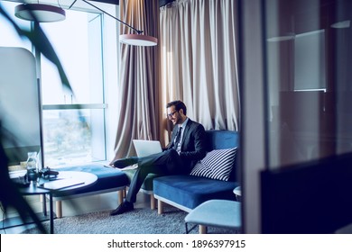 Good-looking Caucasian bearded businessman in suit and with eyeglasses sitting on sofa in his office and using laptop for writing report. - Shutterstock ID 1896397915