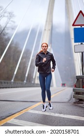 Good-looking caucasian female in sportive clothes running in urban area on bridge, young redhead female in good physique shape engaged in sport fitness, outdoors, on fresh air at spring morning
