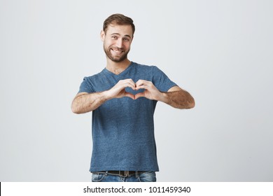 Good-looking Caucasian Male With Beard Holding Hands In Shape Of Heart, Symbolizing Love, Peace And Unity. Smiling Man In Casual Clothes Showing Heart-shaped Hand Gesture Expressing His Affection