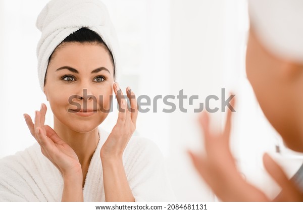 Good-looking\
caucasian middle-aged woman in turban and spa bathrobe after taking\
shower looking at the mirror while applying beauty creme\
moisturizer for anti-age anti-wrinkle\
effect