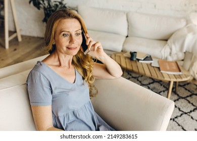 Good-looking charming elderly red-haired lady in blue dress sitting on couch in light stylish reception room, talking on mobile phone with her grandchildren while waiting therapist to come