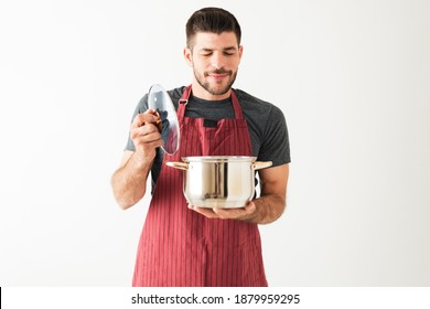 Good-looking Latin Young Man With An Apron Is Carrying A Cooking Pot And Is Smelling The Food He Just Cooked 