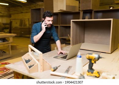 Good-looking Male Carpenter Talking On The Phone With A Customer Inside A Woodshop. Hispanic Man Working On A Carpentry Project On His Laptop 