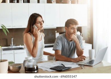 Good-looking Man With Bristle Paying Utility Bills Online On Laptop And His Wife Who Communicates With Her Best Friend Over Mobile Phone Being At Home Kitchen, Working With Documents Of Their Company