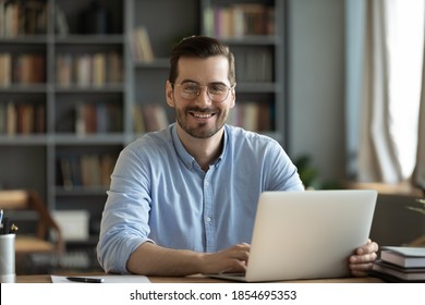 Good-looking millennial office employee in glasses sitting at desk in front of laptop smiling looking at camera. Successful worker, career advance and opportunity, owner of prosperous business concept