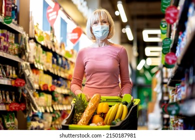 Good-looking Senior Woman In Medical Face Mask Doing Grocery Alone At Supermarket, Female Customer Standing By Shelves With Organic Food With Shopping Trolley Full Of Products, Copy Space