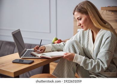 Good-looking woman in a dressing gown writing down notes into her diary on a laptop