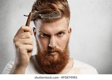 Good-looking young man with red beard holding barbershop accessory. Caucasian barber demonstrating sharp blade of his old-fashioned straight razor, determined to shave clients. Selective focus
