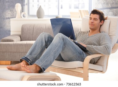 Goodlooking Young Man Relaxing At Home In Armchair, Sitting In Living Room With Laptop Computer, Smiling.?