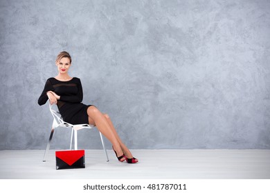 Good-looking Young Woman In A Black Cocktail Dress Sitting On A Gap Chair With Clutch Bag On The Floor