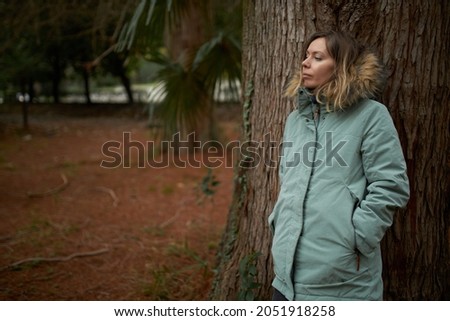 Good-looking young woman in green parka leaning against huge old tree isolated on blurred background of green exotic forest, having pensive face expression, stopping for break during hiking