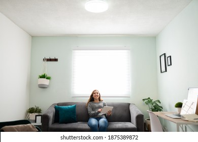 Good-looking young woman sitting on the sofa and turning on a smart light bulb with her tablet  - Shutterstock ID 1875389872