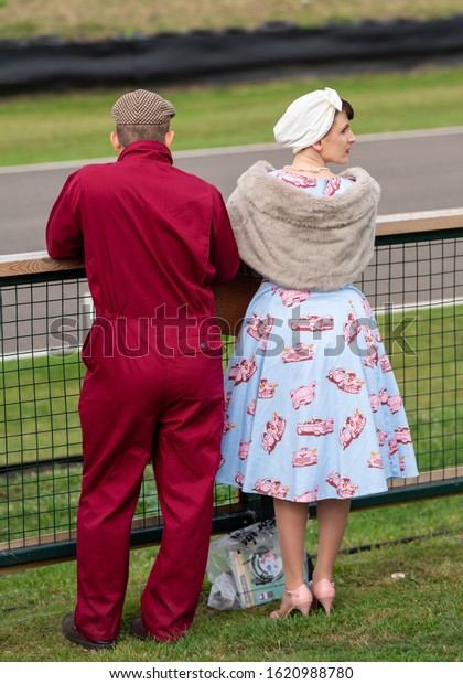 Goodwood, Sussex / UK - 13 September 2019: A couple\
in vintage clothing stand track side at retro Revival. The guy has\
red motorsport overalls, the lady a blue print dress with car motif\
and fur shawl