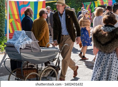 Goodwood, Sussex / UK - 14 Sept 2019: A Man Pushes A Vintage Grey Silver Cross Pram With Large Wheels, Hood And Brown Wicker Picnic Hamper Past People Wearing Vintage Dress At The Revival Event. 