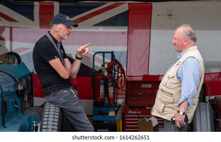 Goodwood, Sussex / UK - 6 July 2019: Two men talk while leaning on vintage cars. One man looks cross and points a finger, the other leans back as if chastised. A large union jack flag is on the wall.